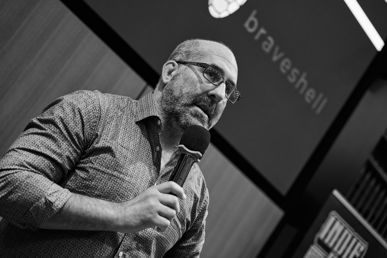 A speaker in glasses holds the mic in front of his Braveshell title slide.