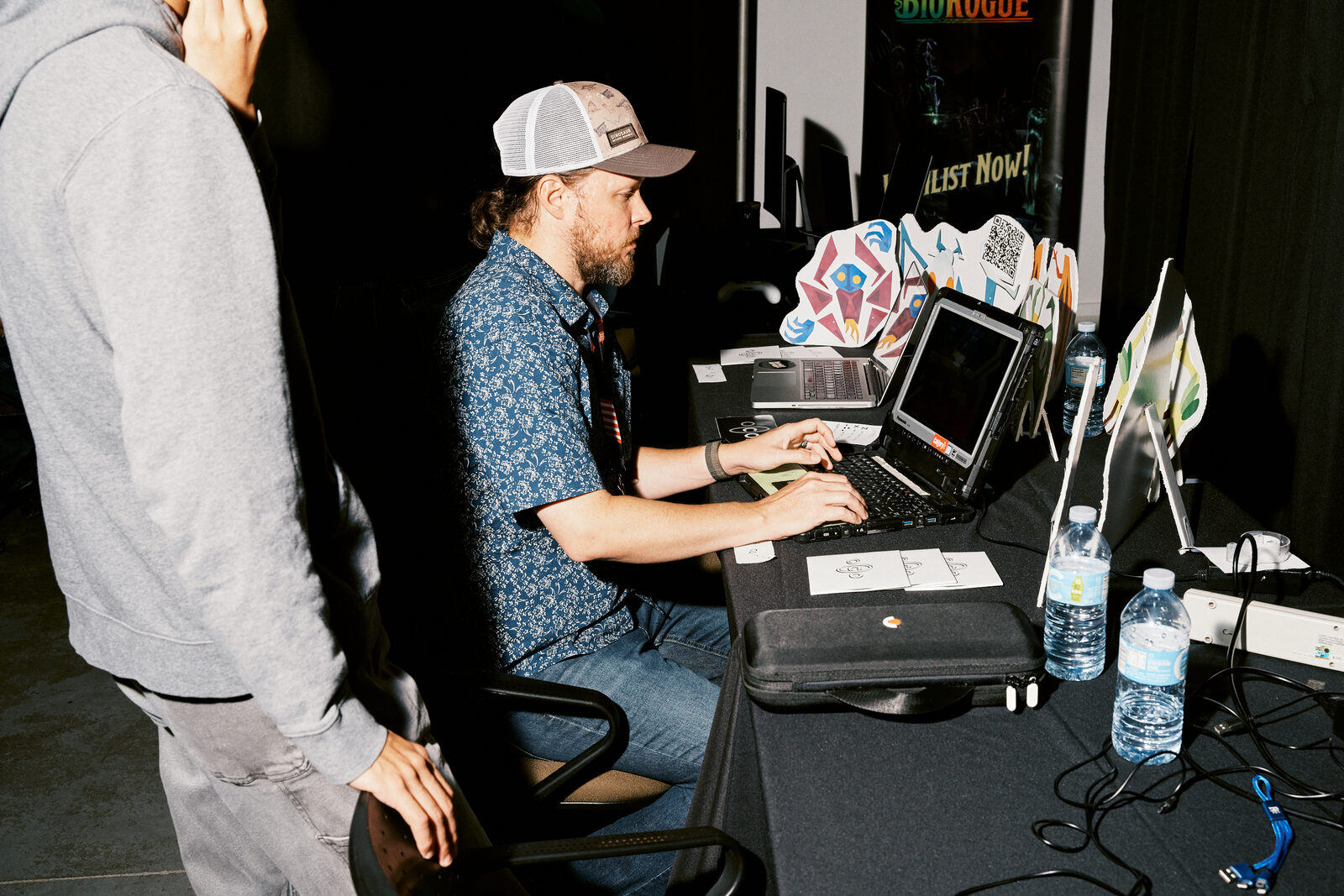 A developer in a trucker cap works at his laptop at the BioRogue booth, sticker standees and water bottles down the table.