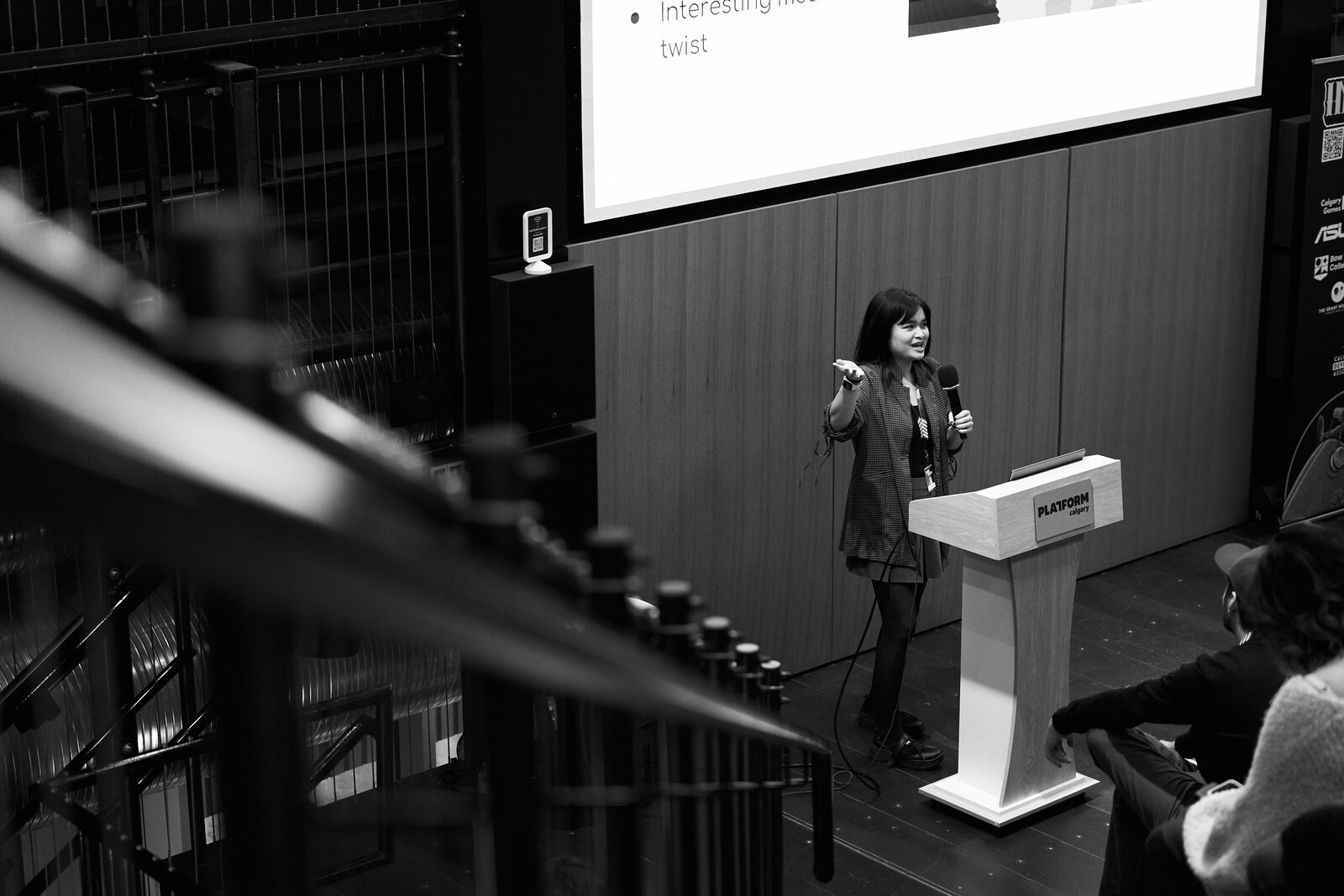 From the balcony, past the stage lighting rig: a speaker alone at the Platform podium with her slide behind her.