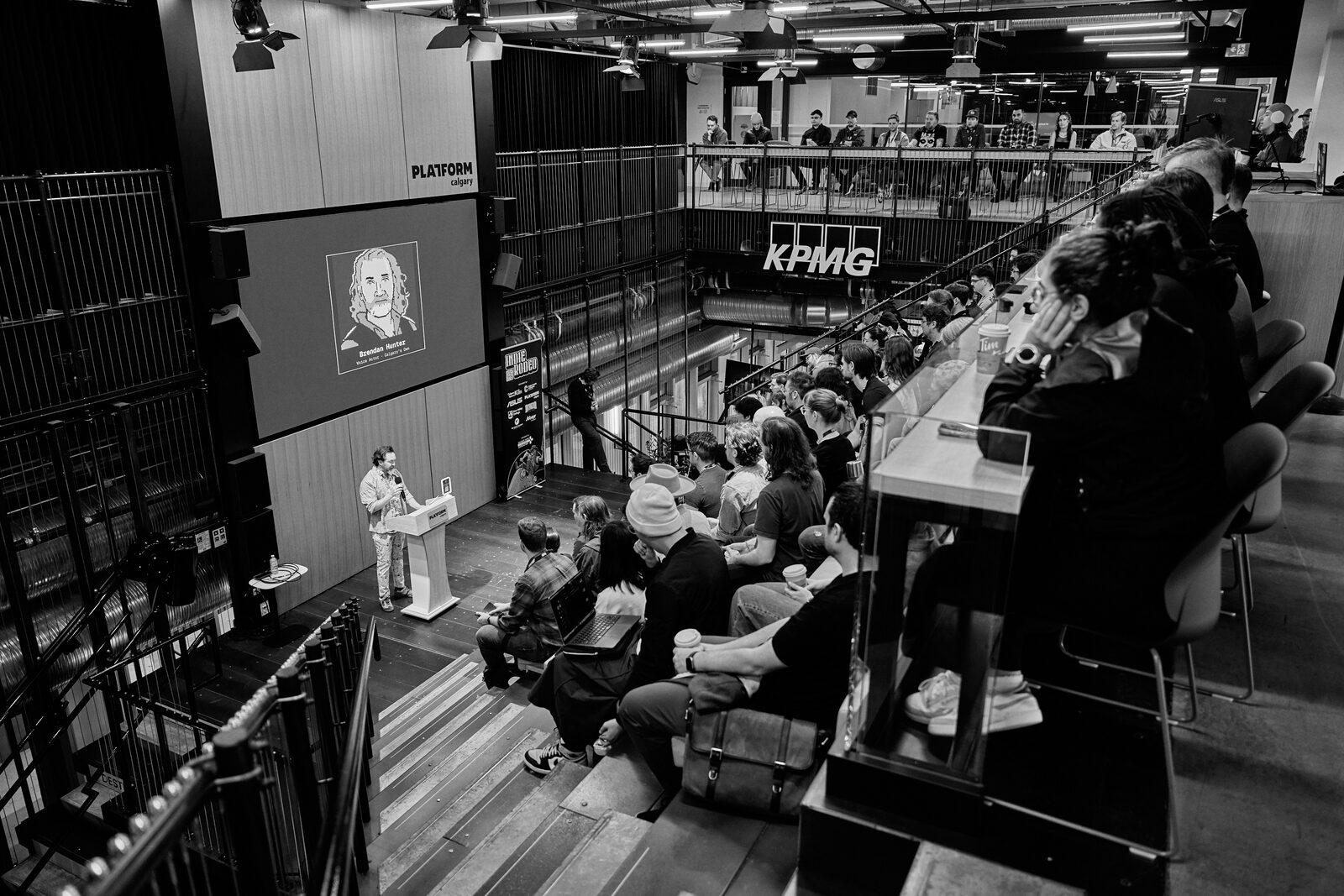 Looking down into the Platform Calgary atrium from the balcony: a speaker at the lit podium, their portrait on the big screen, attendees filling the bleacher steps under the KPMG sign.