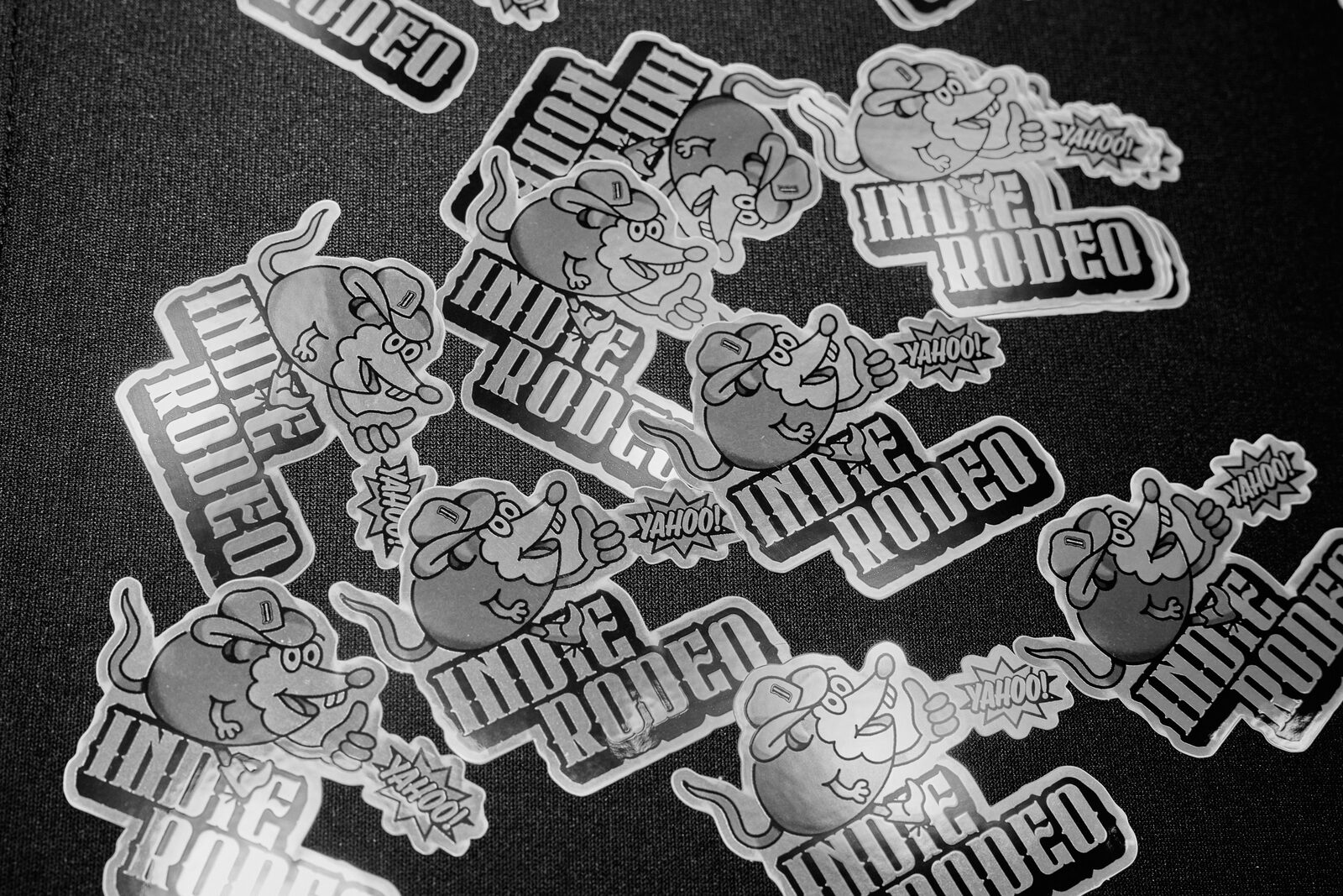 A scattered pile of holographic Indie Rodeo stickers on a black tablecloth, the grinning rat mascot and the wordmark catching the light.