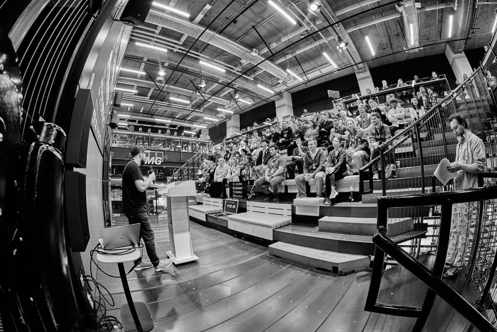 Fisheye view from beside the stage: a speaker at the podium facing a packed house of attendees on the bleachers.