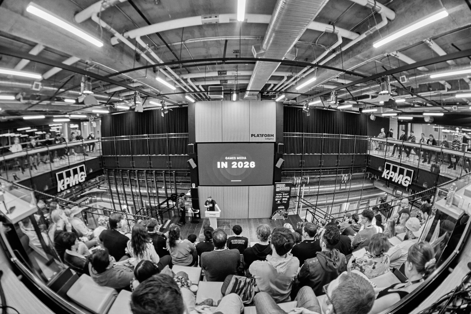 Fisheye view from the back of the room, the full atrium in frame, the stage screen reading "IN 2026" over the seated crowd.