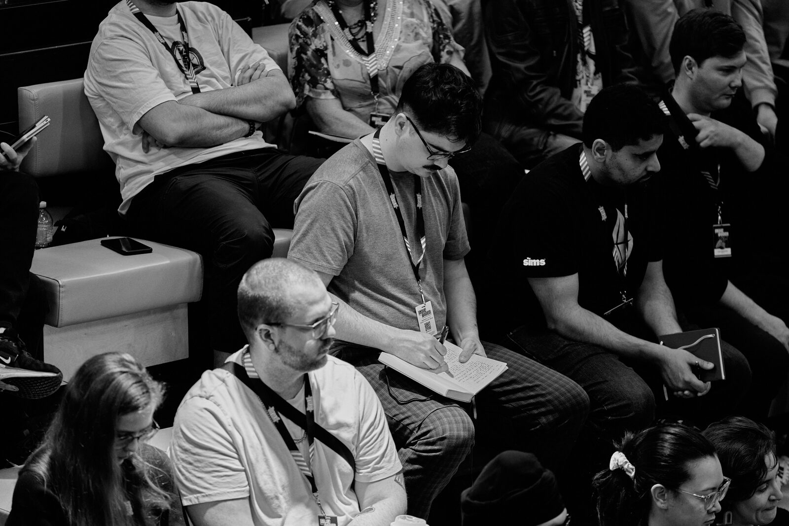 Attendees on the bleachers during a talk; one of them is taking notes in a paper notebook.