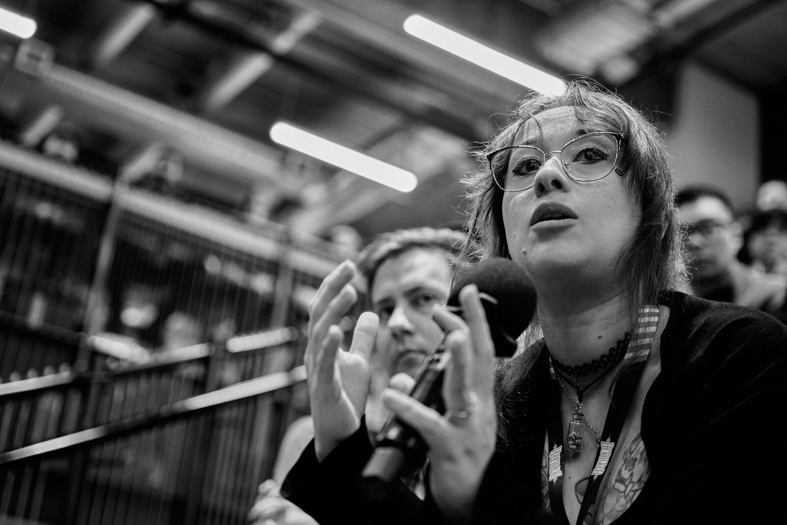 An attendee with glasses gestures as she asks a question, mic in hand, a second attendee listening beside her.