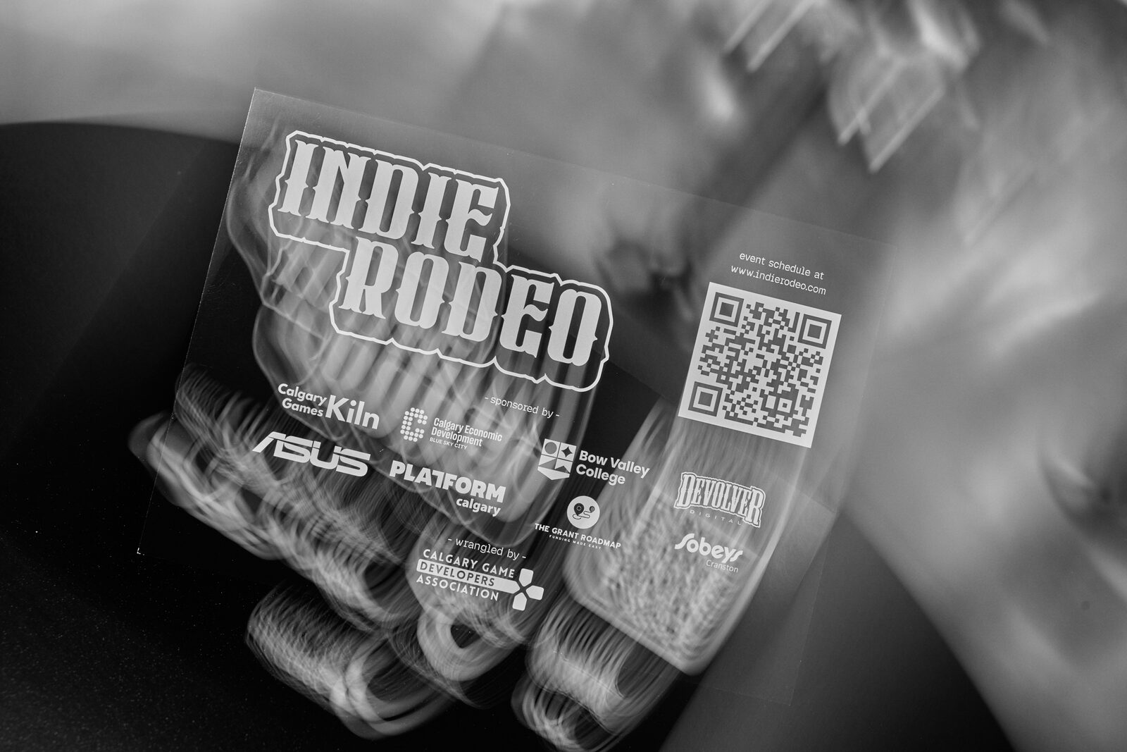 An Indie Rodeo badge and lanyard close up, the QR square and the sponsor row legible while the background falls away into bokeh.