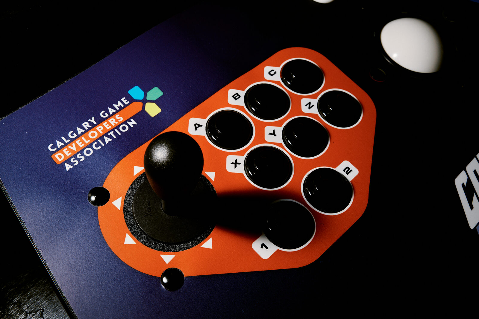 A Calgary Game Developers Association arcade stick, orange faceplate and black ball top, resting on a desk under a bare bulb.