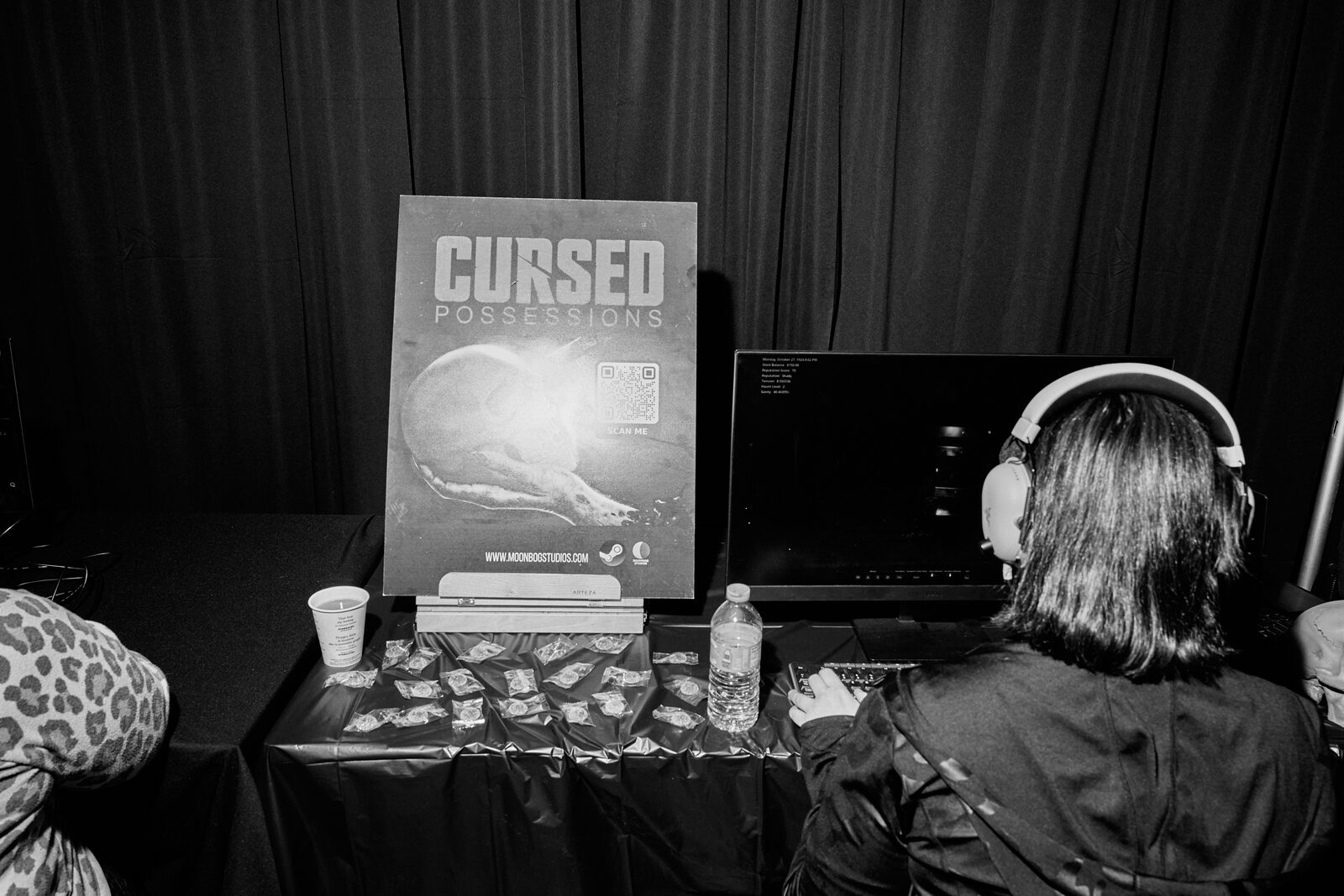 A showcase booth for Cursed Possessions — pull-up banner beside a monitor, a player in headphones at the keyboard.