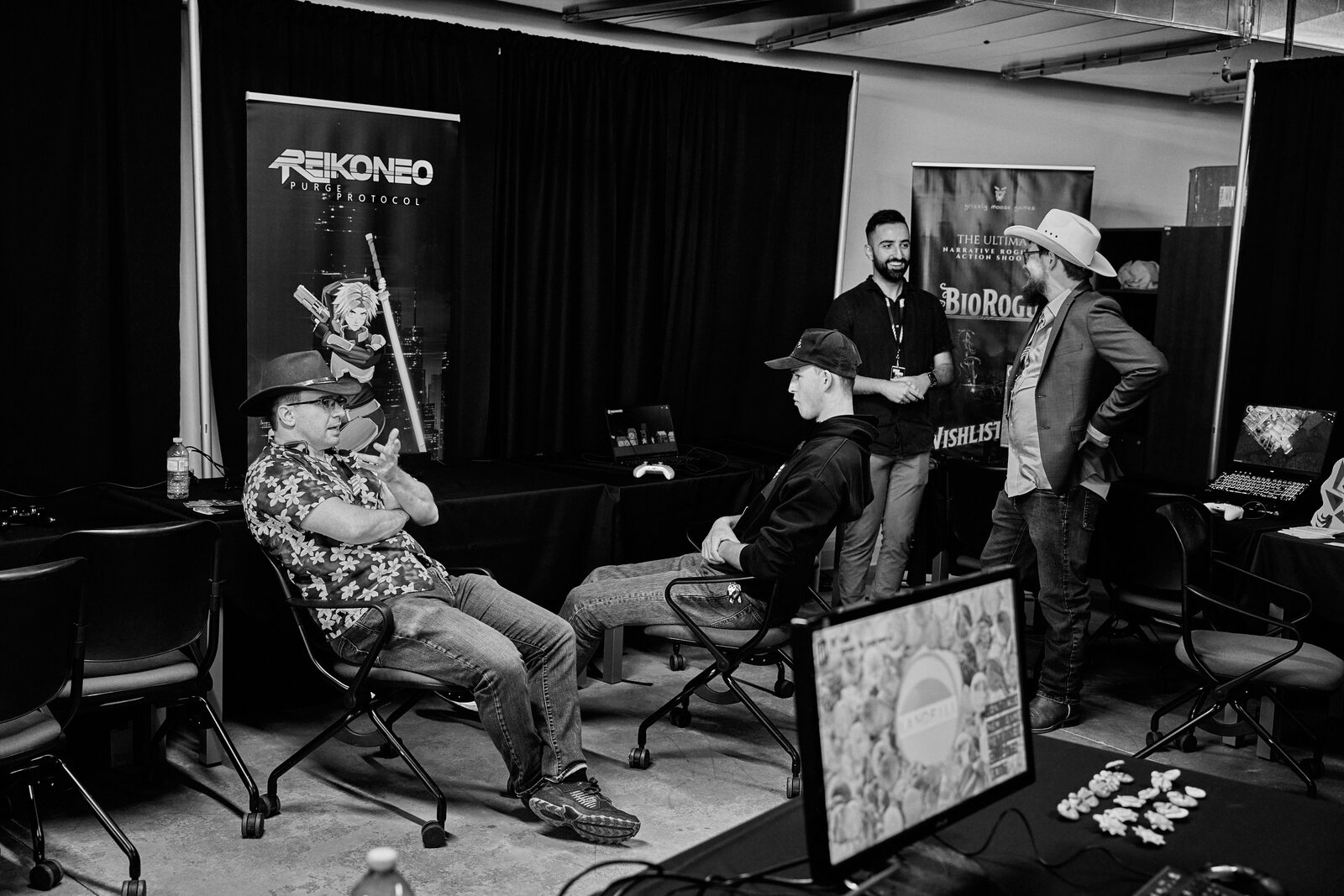 Four attendees in cowboy hats and caps talking between showcase stations, Reikoneo and BioRogue banners behind them.