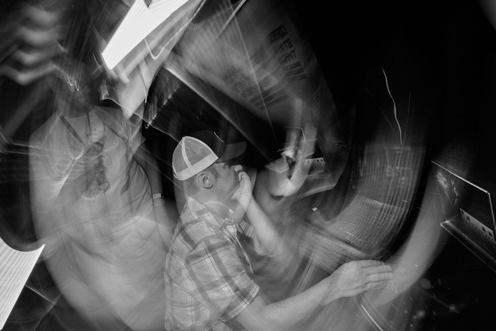 A spun long-exposure of the showcase floor: an attendee in a cap sharp at the centre while the booths smear into streaks around him.