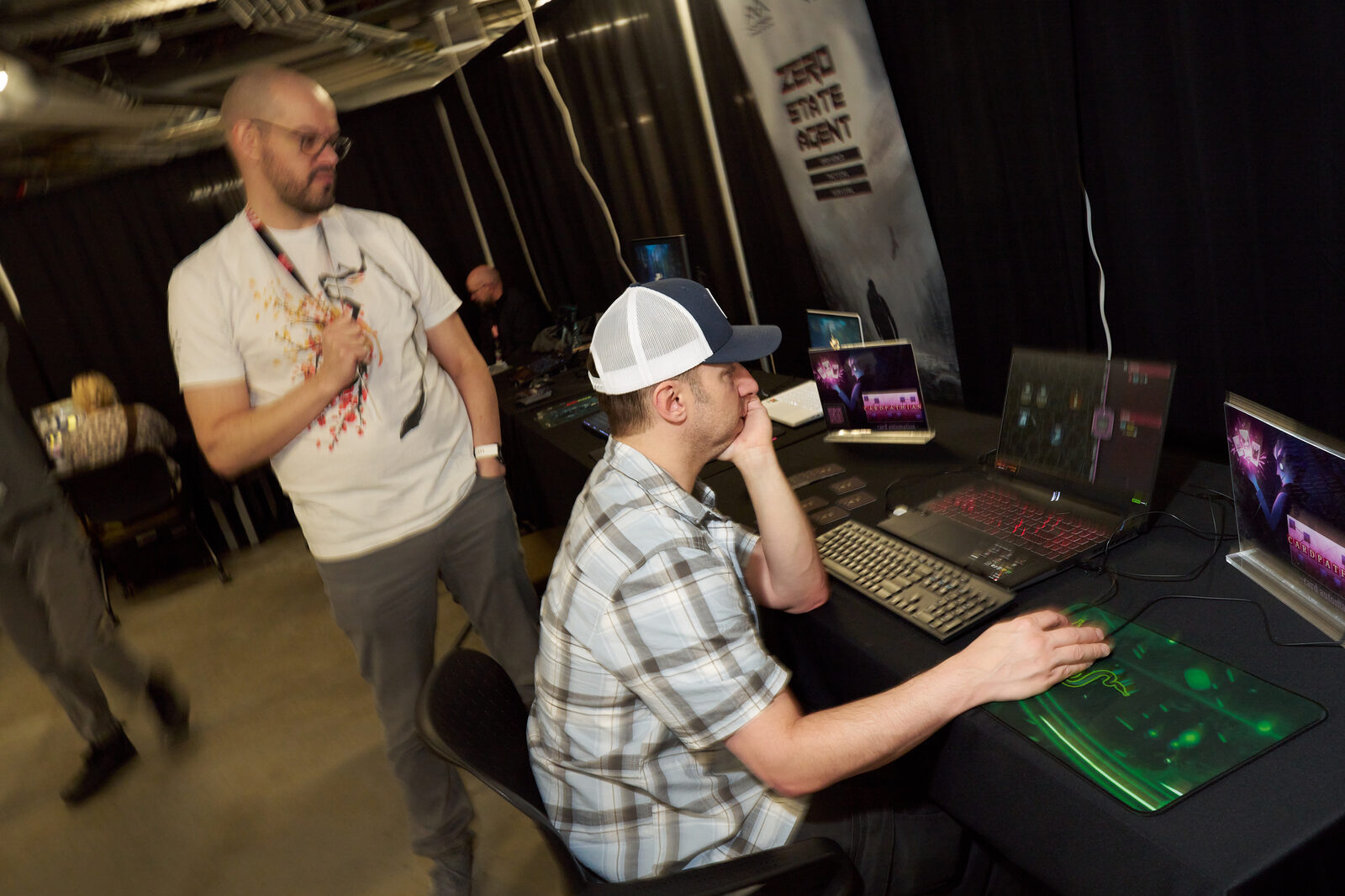 Two developers at a showcase station, one seated at the keyboard mid-demo, the Zero State Agent banner behind them.