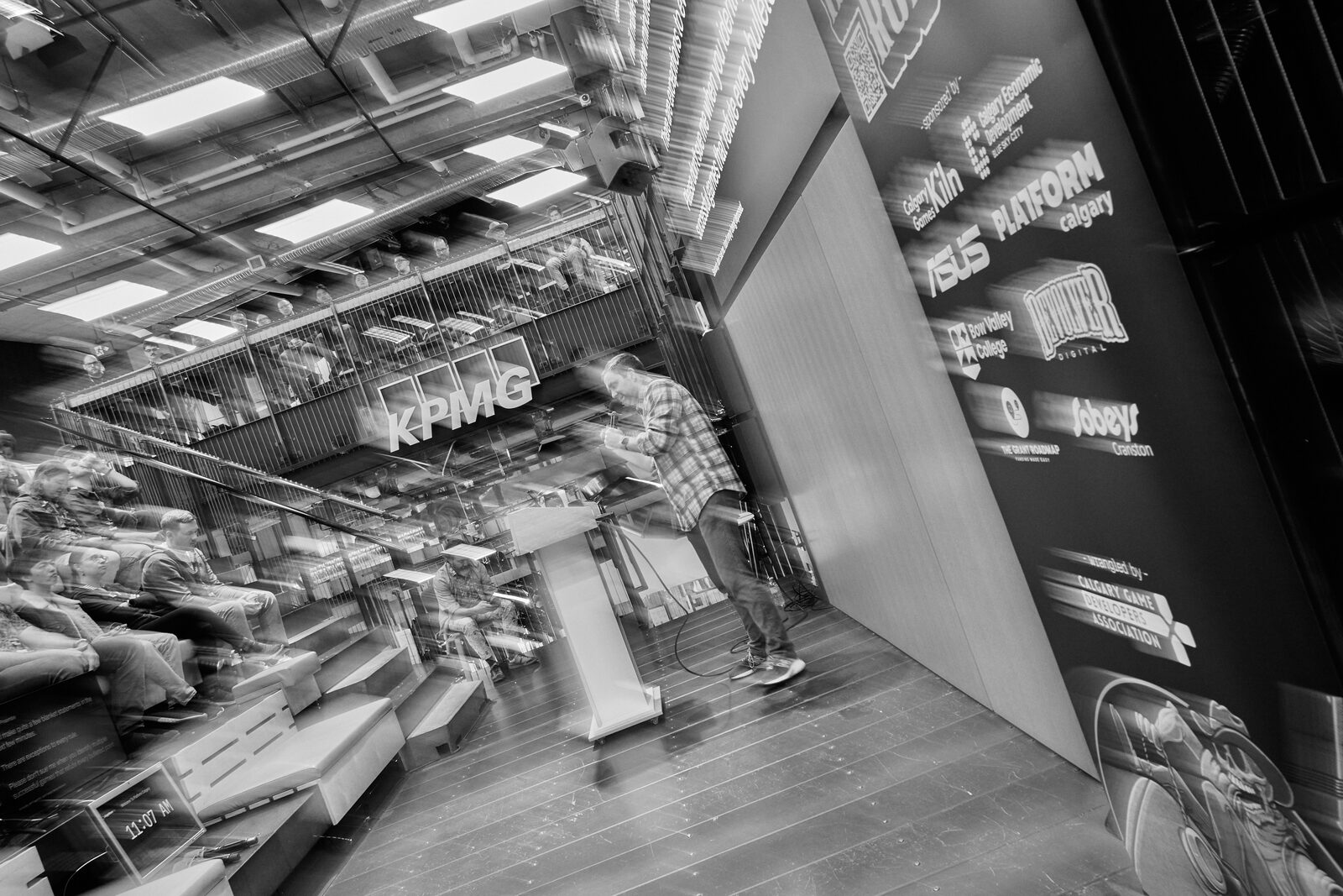 A motion-blurred wide of the atrium: the speaker at the podium held steady while the room swings past, sponsor wall to the right.