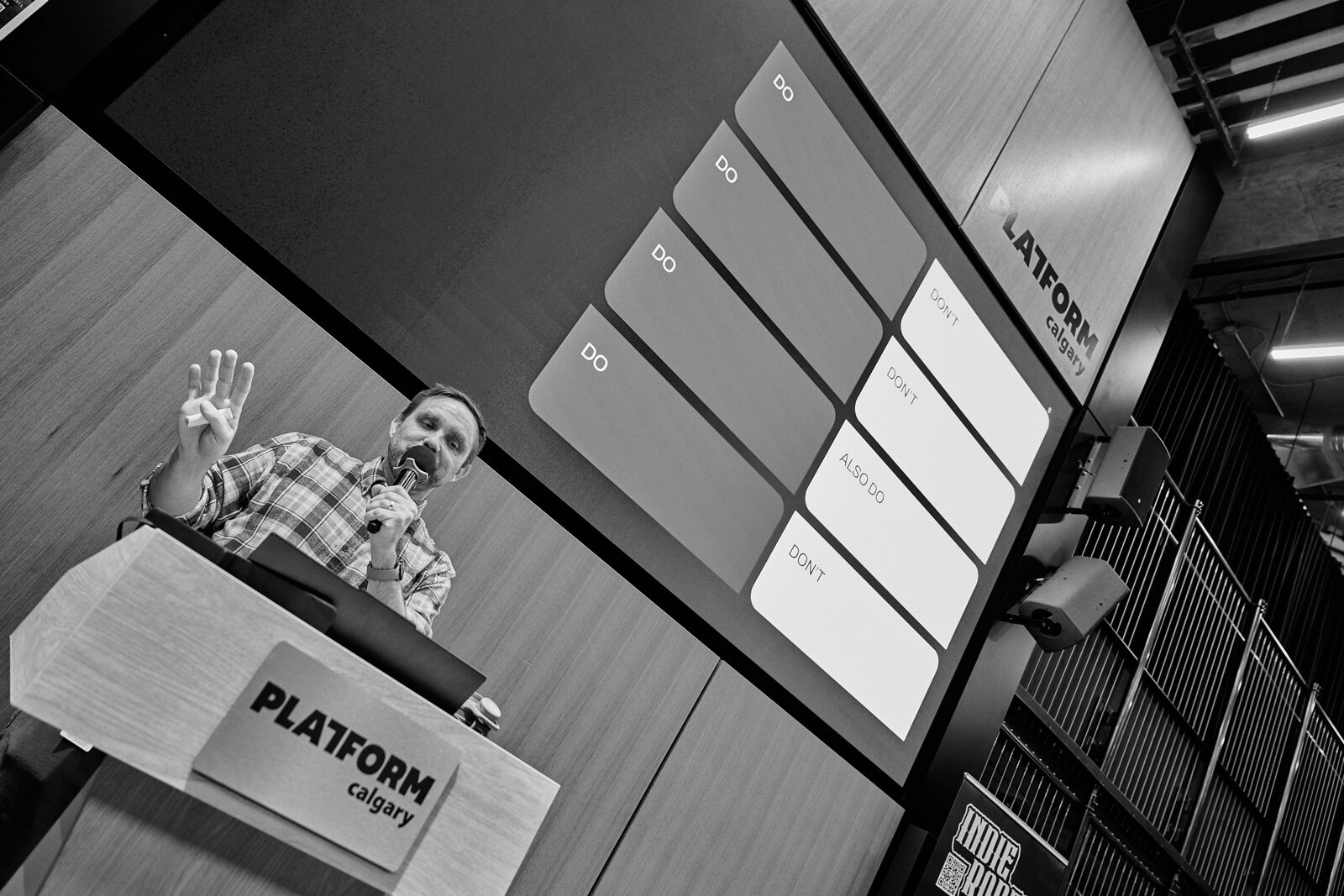 A speaker at the Platform Calgary podium with a hand up mid-point, their slide grid filling the screen behind.