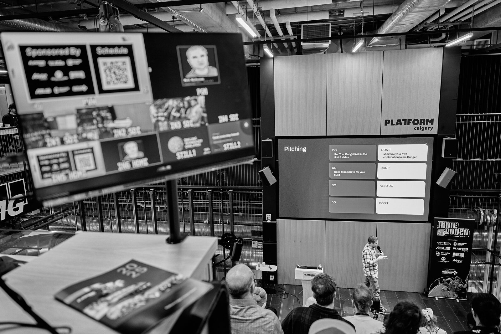 The atrium from the side: a "Pitching" slide on the main screen, the run-of-show and sponsor grid on a second monitor, the speaker small on stage.