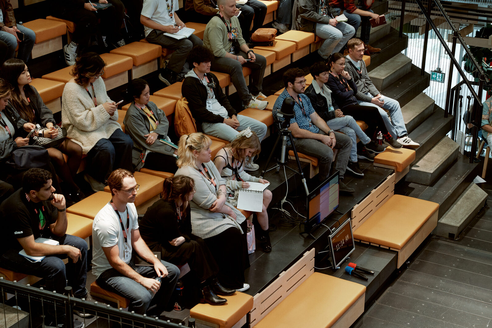 Attendees spread across the orange bleacher seats during a talk, laptops open, a few turned toward each other mid-conversation.