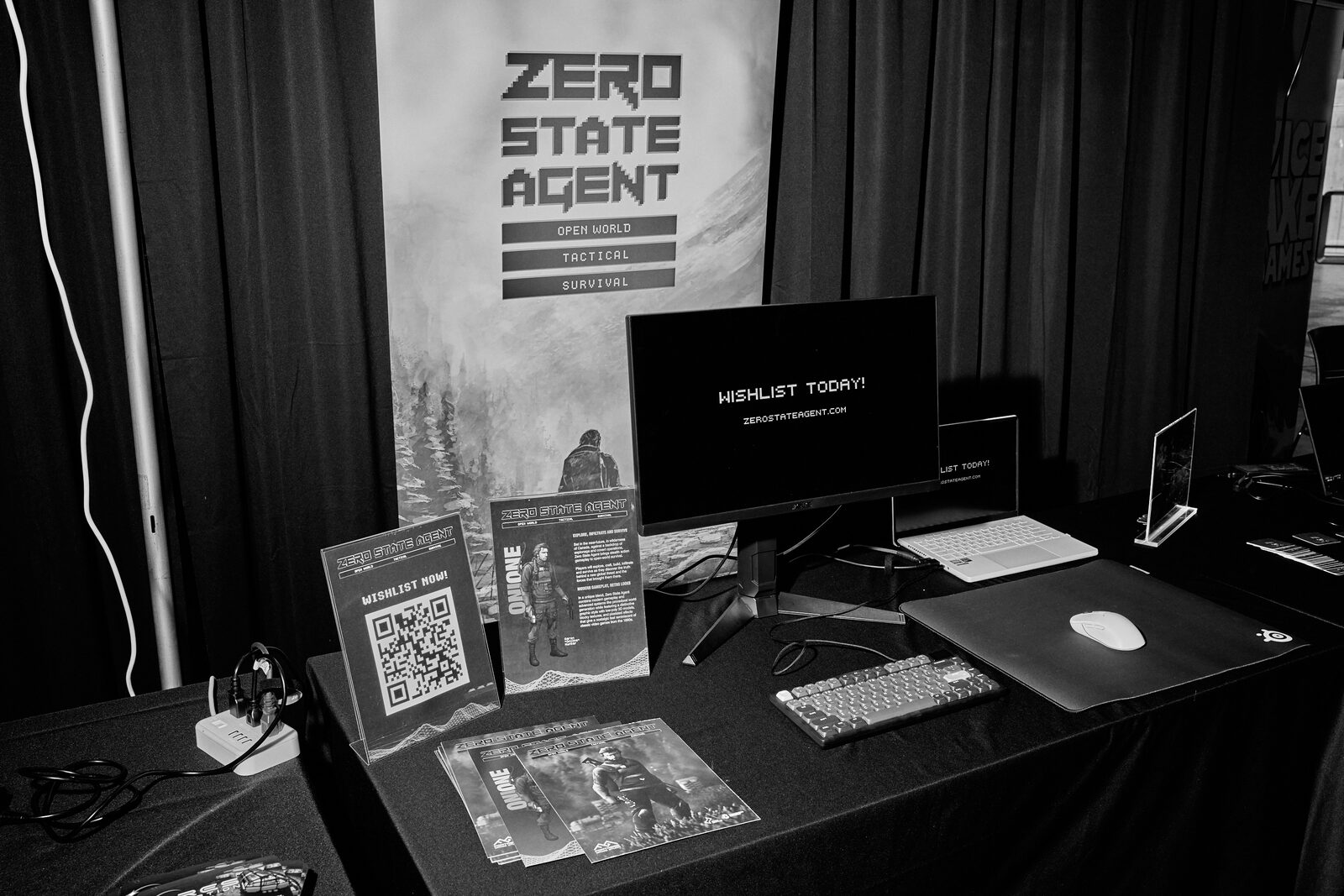 The Zero State Agent booth: banner, a monitor reading "Wishlist today!", QR cards, keyboard and mouse on black cloth.