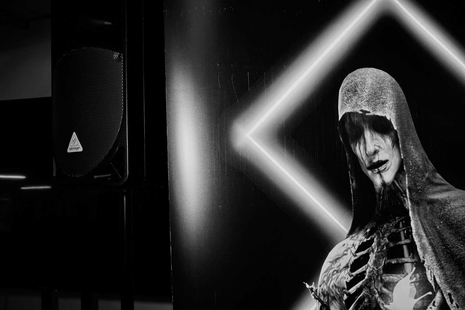 A hooded figure in skeletal makeup stands lit from behind by a neon chevron, the darkest frame of the day.