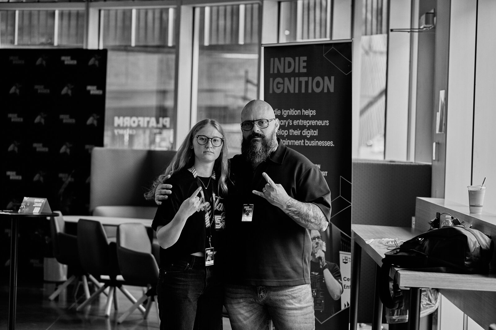 Two attendees throwing metal horns for the camera in front of the Indie Ignition banner.