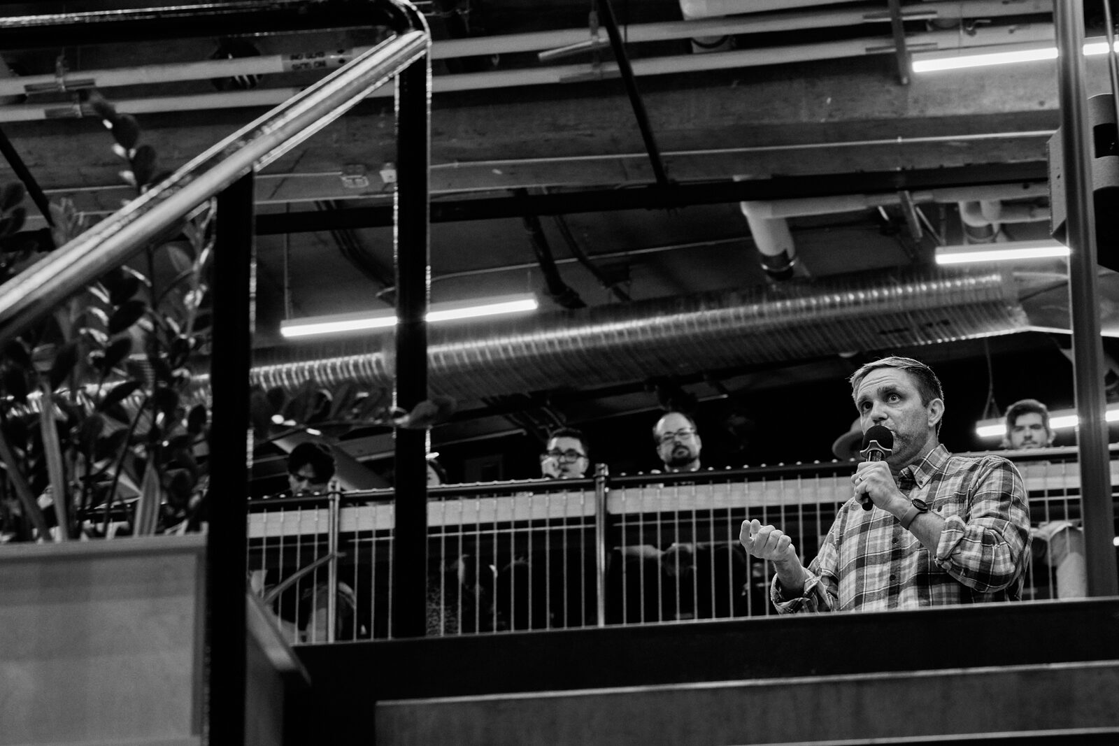 A speaker with a mic seen past the atrium railing, attendees watching from behind the mesh barrier.