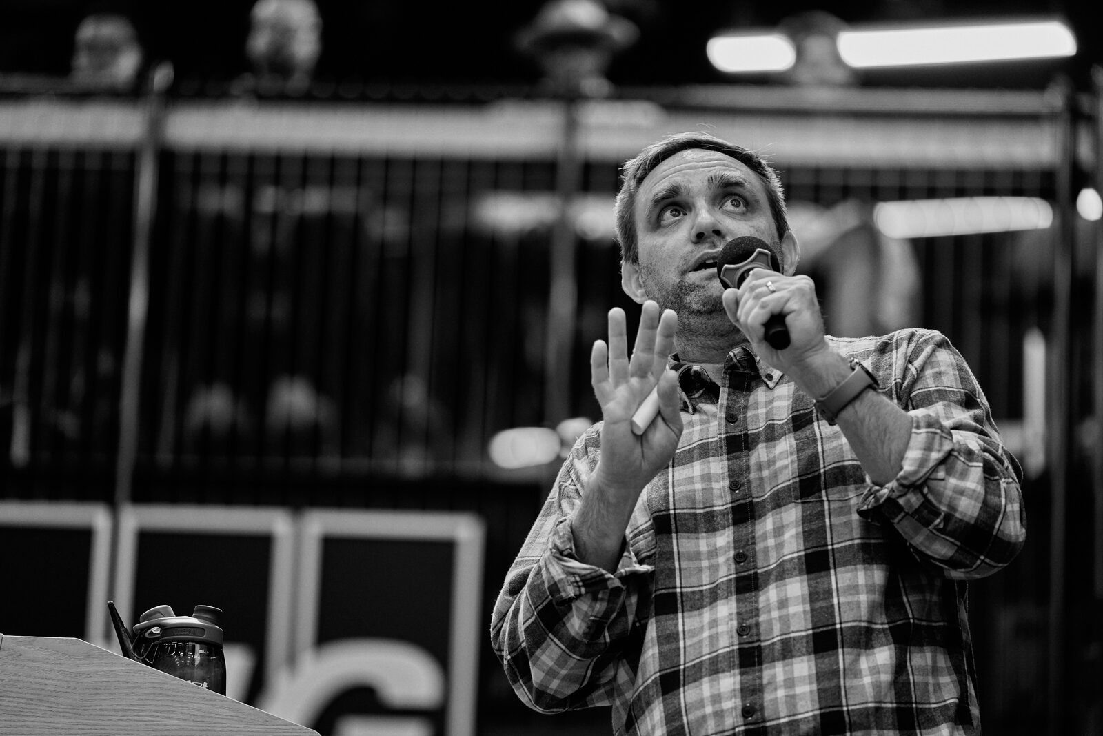 A speaker in a beanie holds the mic close and lifts both hands mid-point, the KPMG wall out of focus behind him.