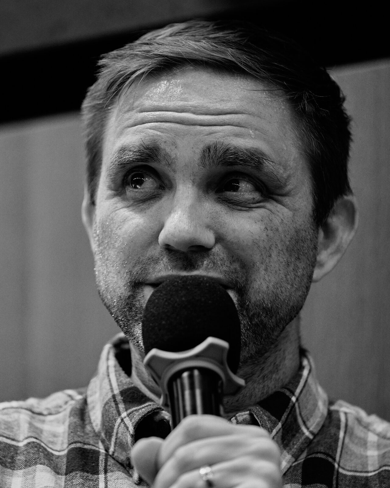 Tight portrait of a speaker in a plaid shirt holding the mic close, caught mid-thought.