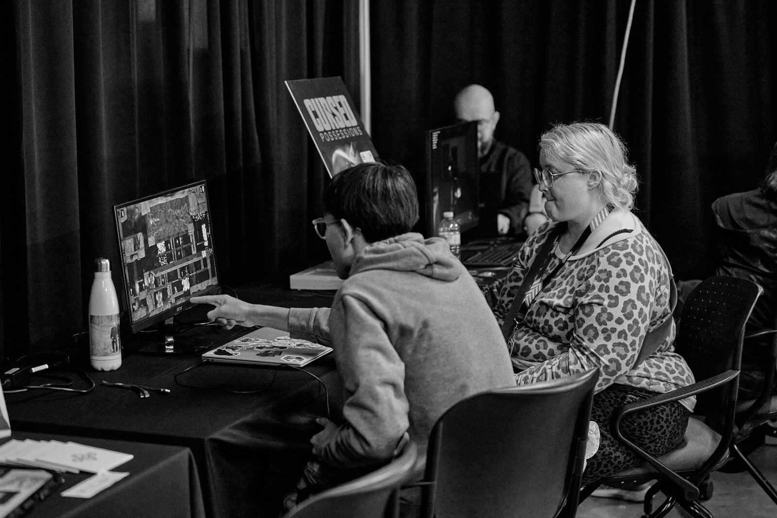 Two attendees at a showcase monitor, one pointing something out on screen, the Cursed Possessions banner beside them.
