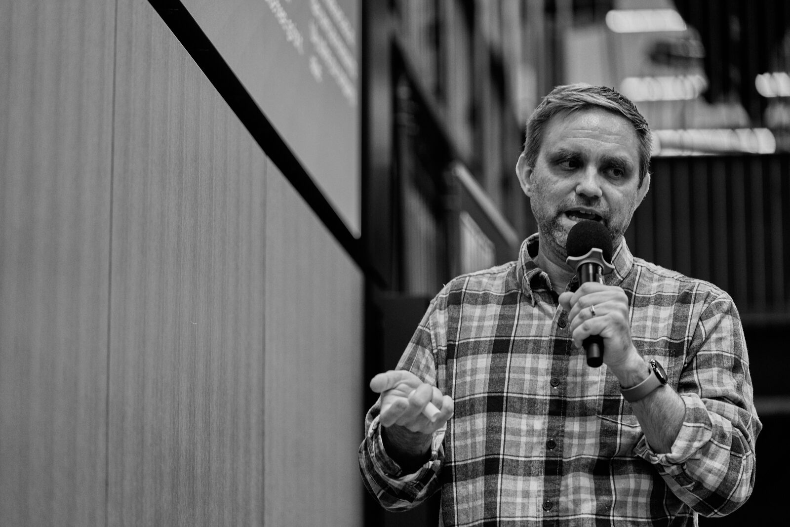 A speaker in a plaid shirt works the mic with his free hand raised, his slide glowing behind him.