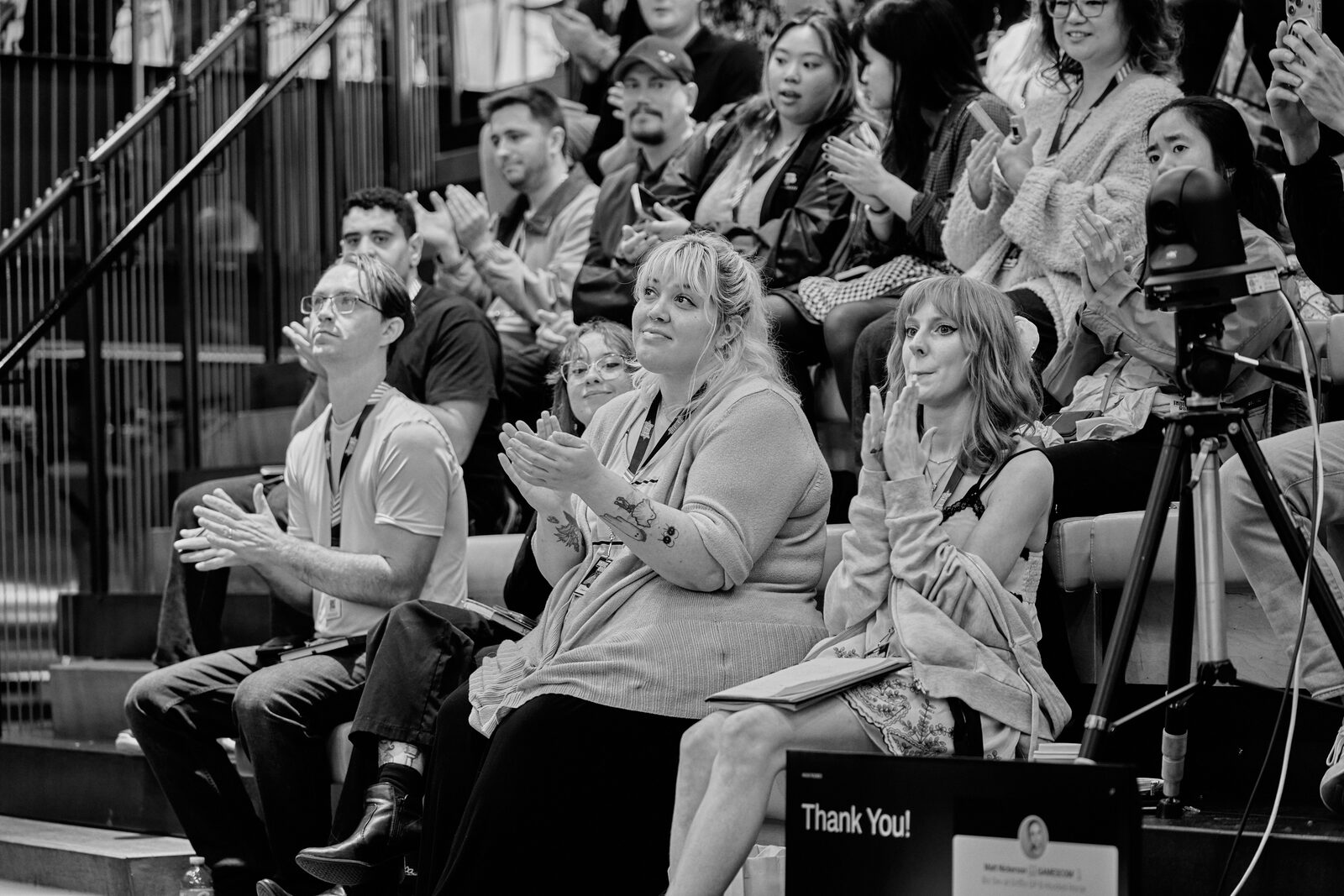 A bleacher row mid-applause, a dozen attendees clapping, a camera on a tripod at the end of the row.