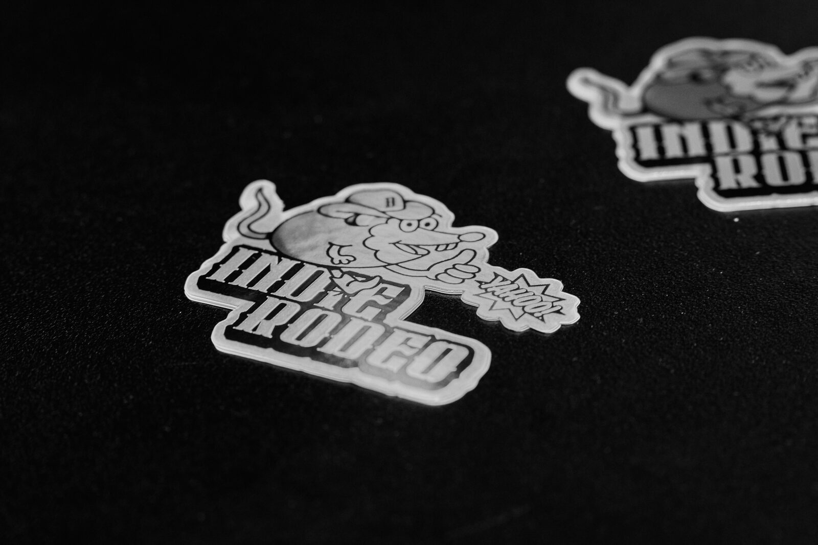 Two holographic Indie Rodeo stickers on a black surface, the rat mascot glinting under a hard light.