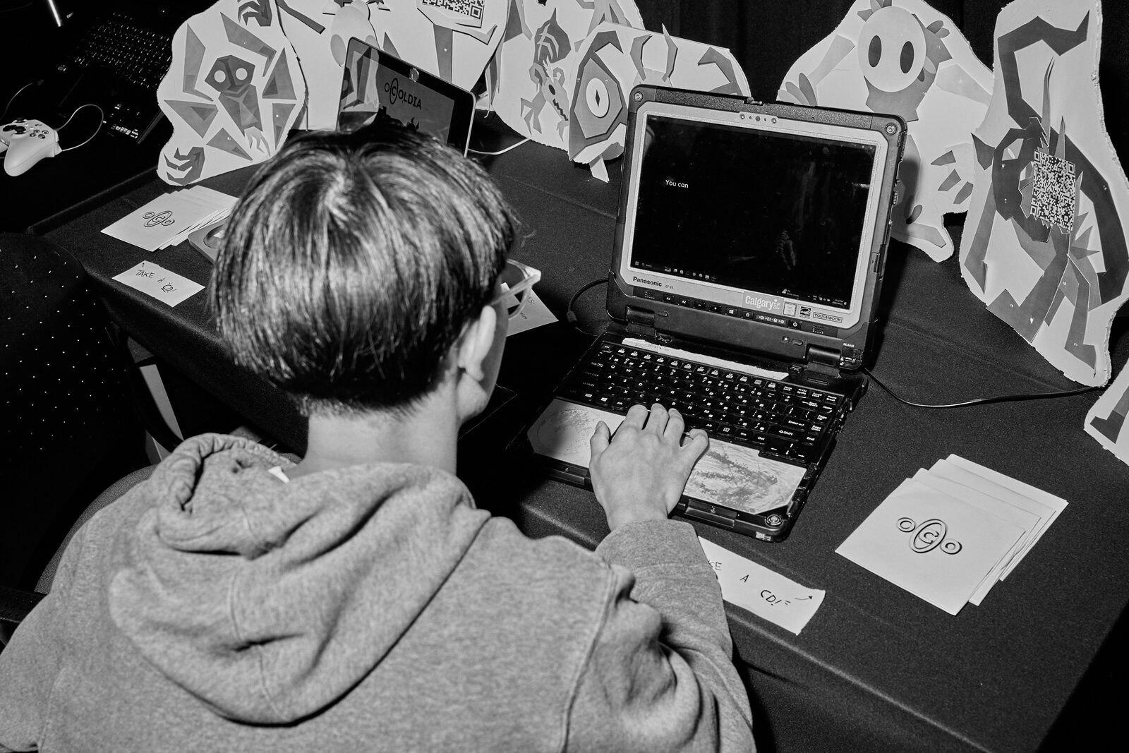 An attendee at a laptop on the showcase floor, ringed by paper standees and cut-out character art.