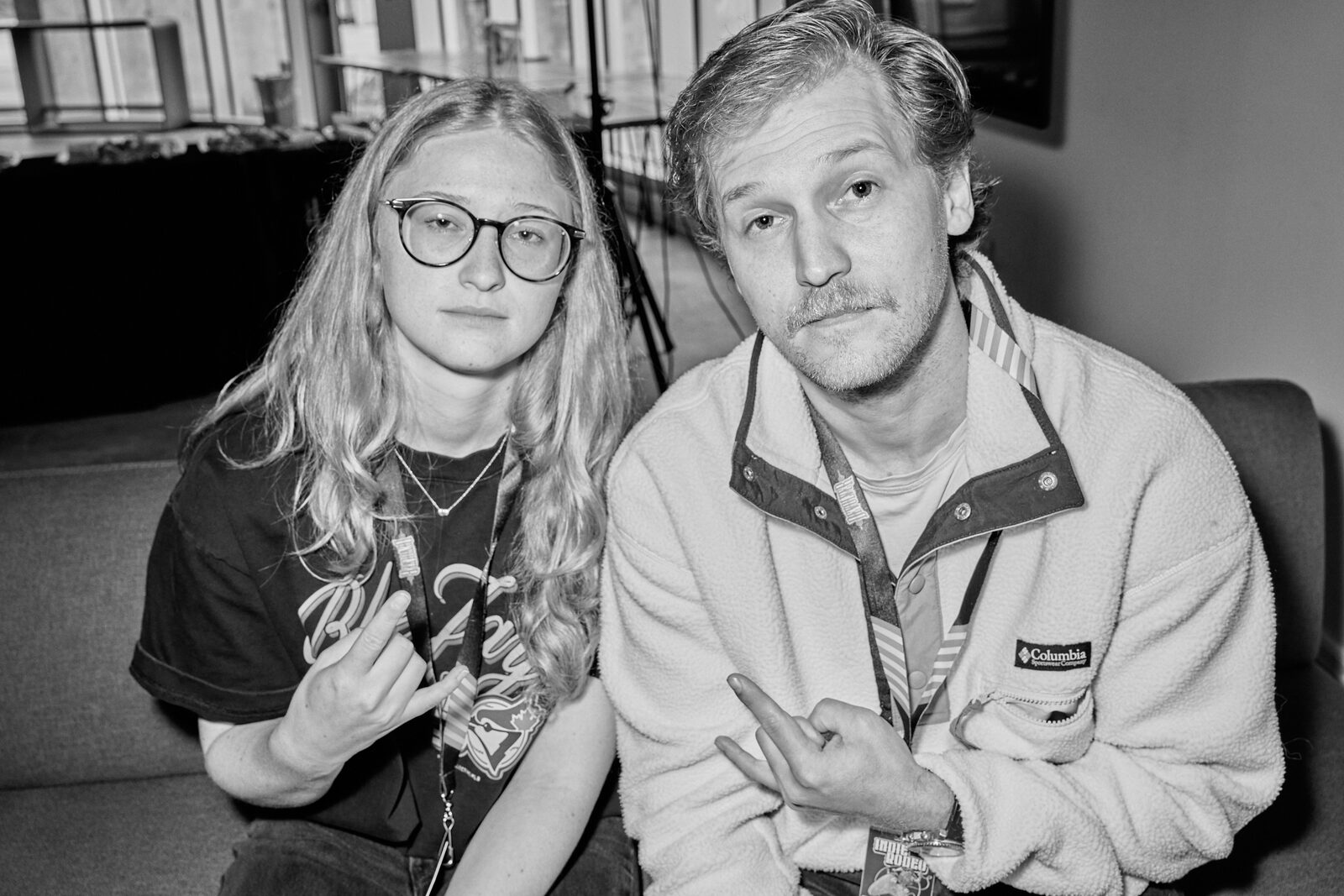 Two attendees side by side on a couch, one in glasses and a band tee, the other in a fleece, both throwing a hand sign at the camera.