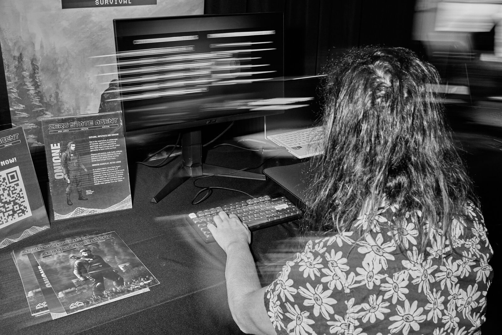An attendee plays a demo at a showcase station, the monitor blurred with motion, flyers and a QR card on the table.