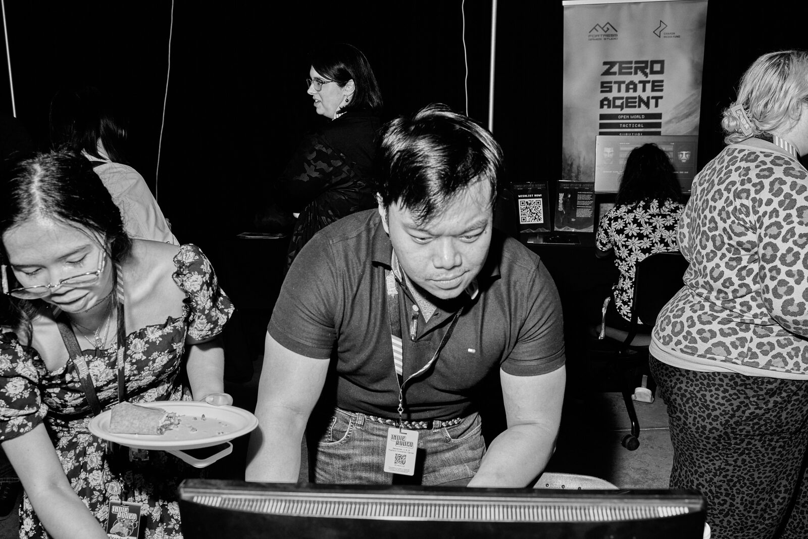 An attendee carries a plate of food past the showcase monitors while another leans in to play.