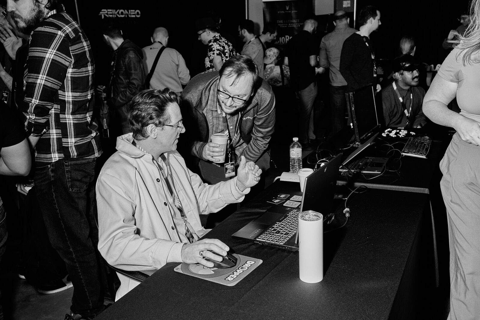Two attendees at a showcase station on the crowded floor, one at the mouse, the other leaning in with a drink.
