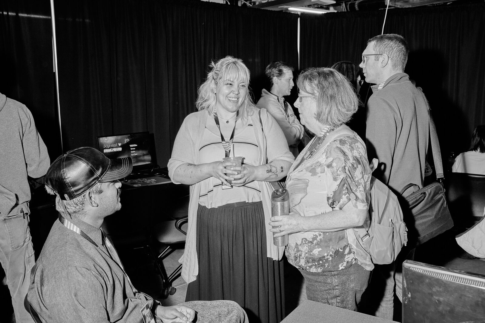 Attendees standing with drinks mid-conversation on the showcase floor, a seated attendee in a cowboy hat in the foreground.