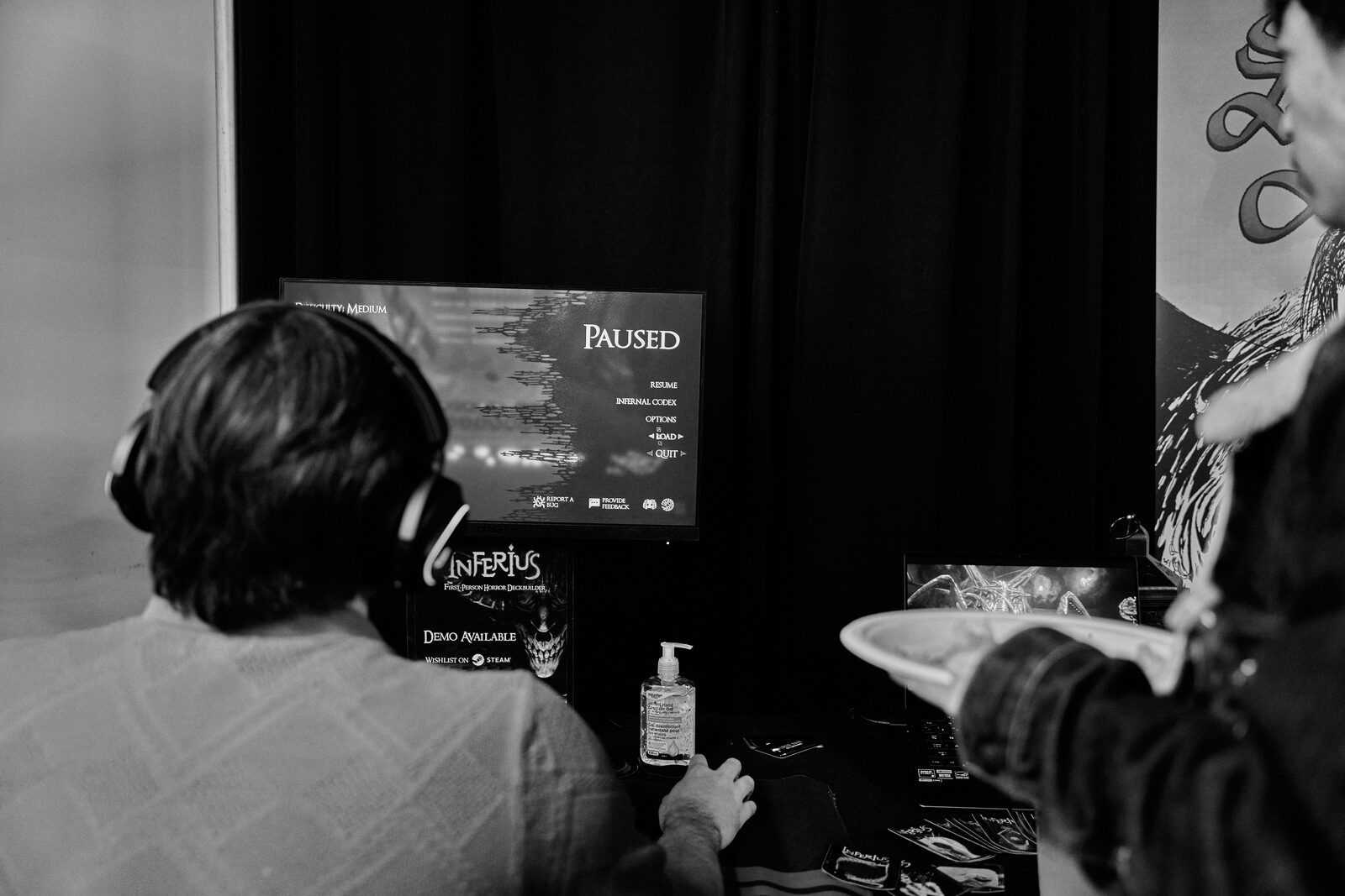 A player in headphones at the INFERIUS station, the game paused on screen, another attendee waiting beside him with a plate.