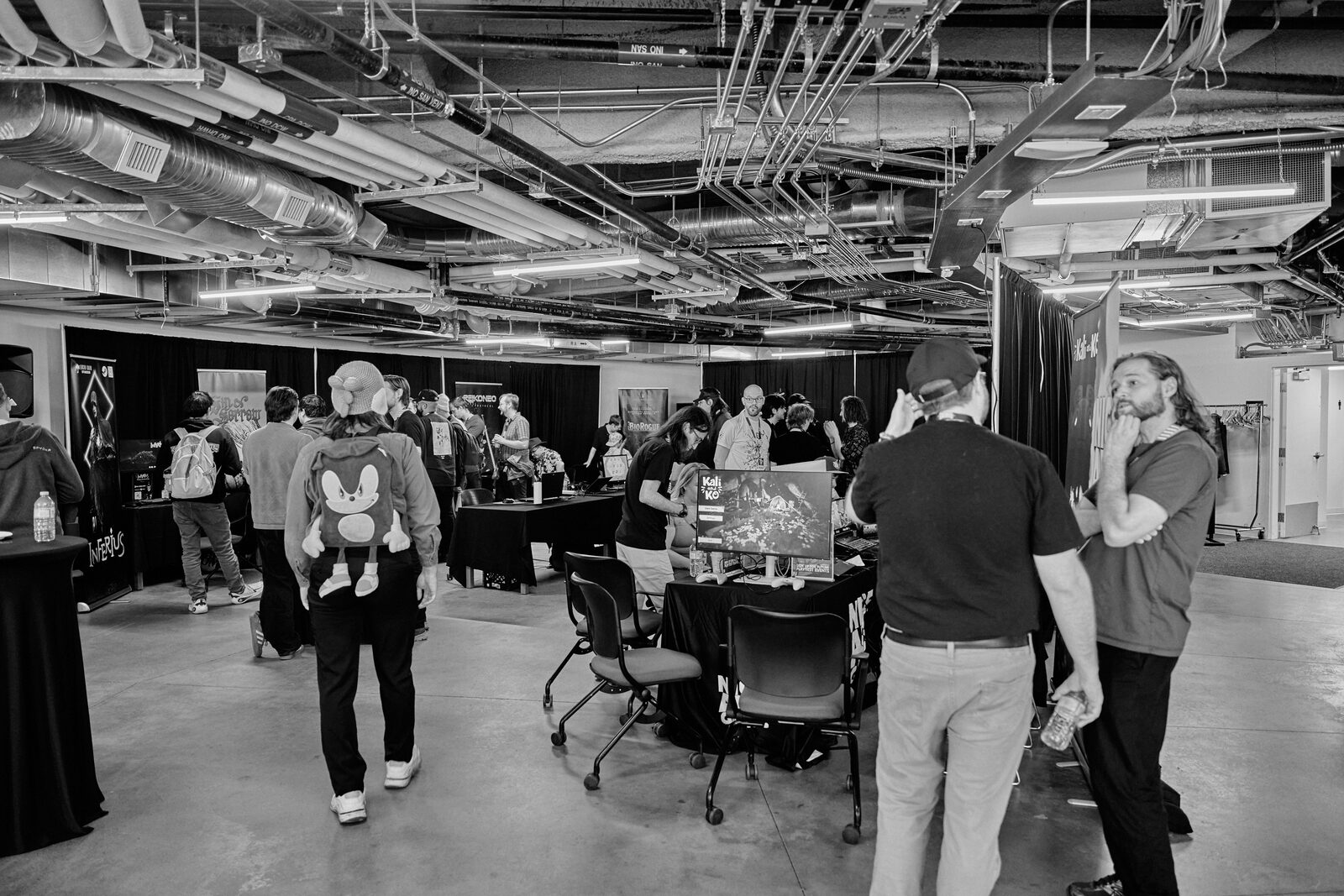 Wide of the showcase room under its exposed ceiling: attendees moving between stations, one with a Sonic backpack.