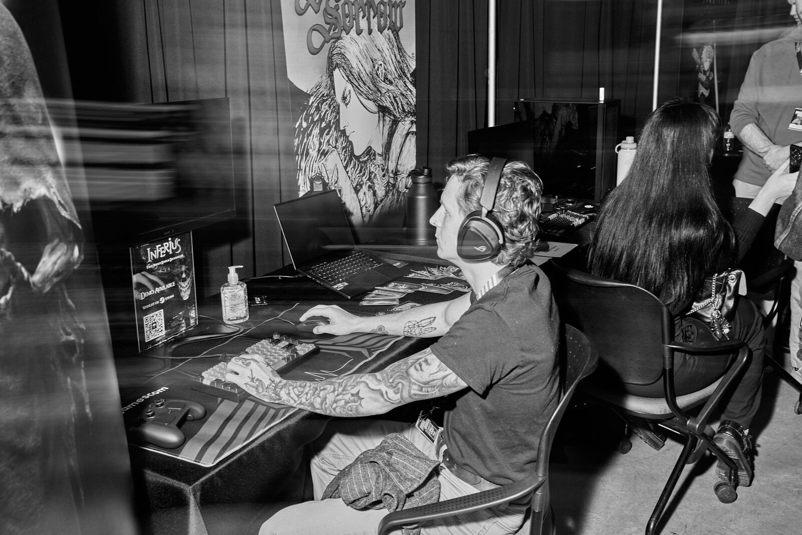 A player in headphones at a keyboard, tattooed arms on the desk, a large illustrated poster over the station.
