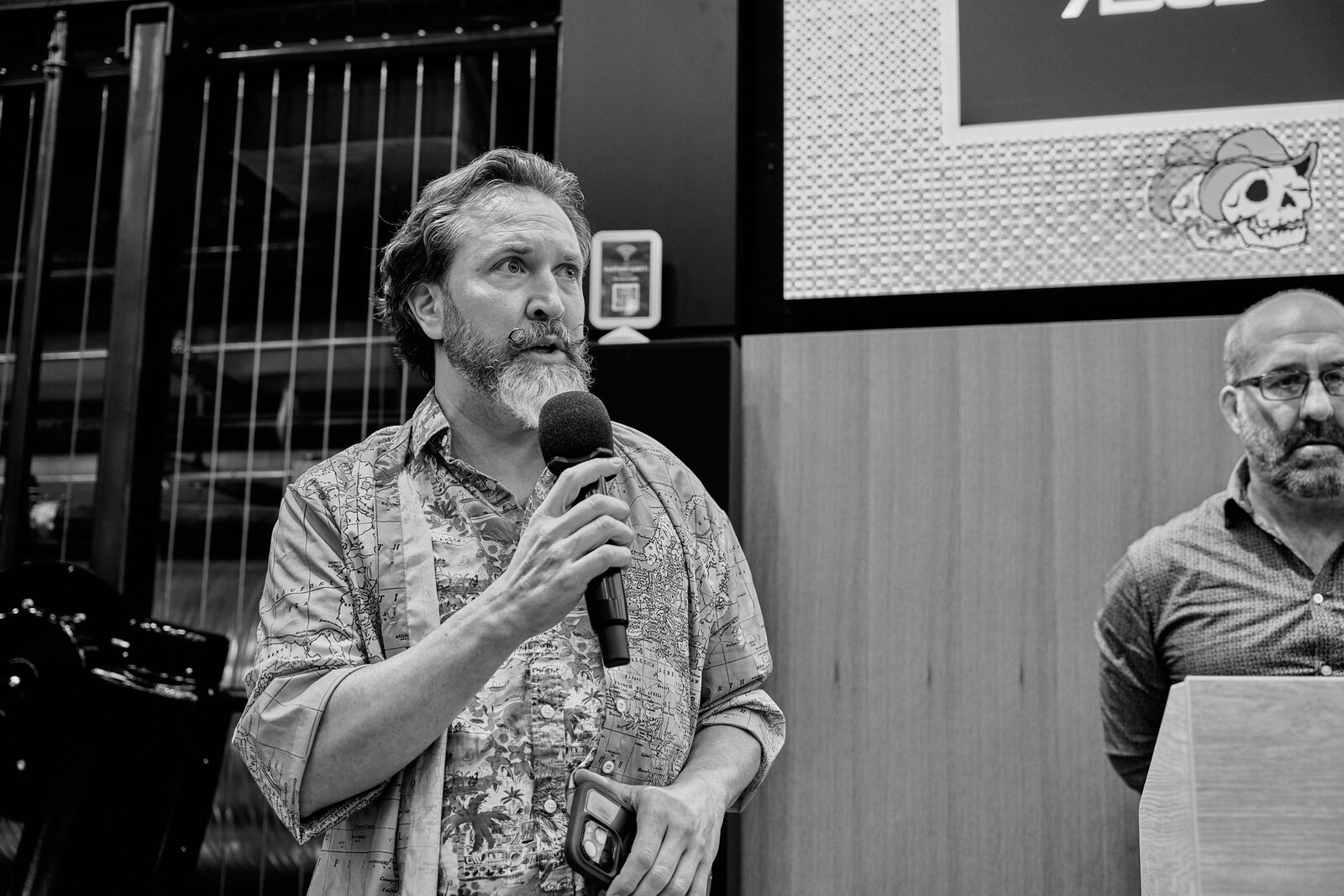 A speaker in a patterned shirt talks into a handheld mic, a second presenter at the lectern to his right.