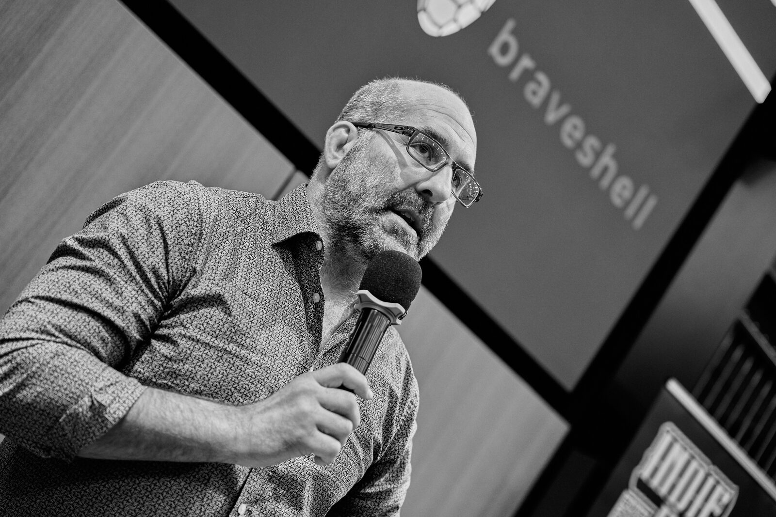 A speaker in glasses holds the mic in front of his Braveshell title slide.