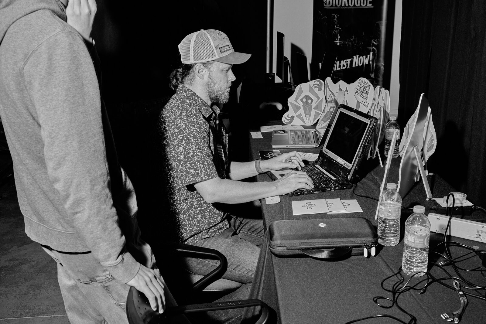A developer in a trucker cap works at his laptop at the BioRogue booth, sticker standees and water bottles down the table.