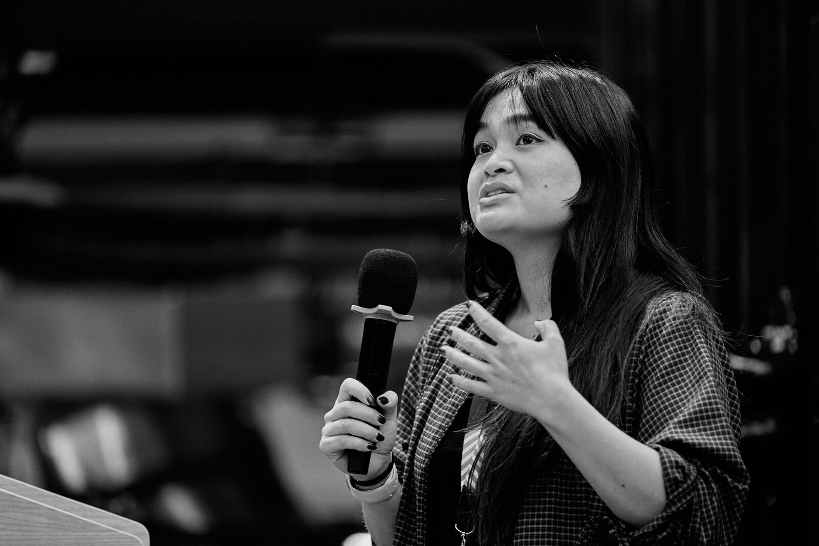 A speaker holds the mic in both hands and looks up mid-sentence, the room dark behind her.