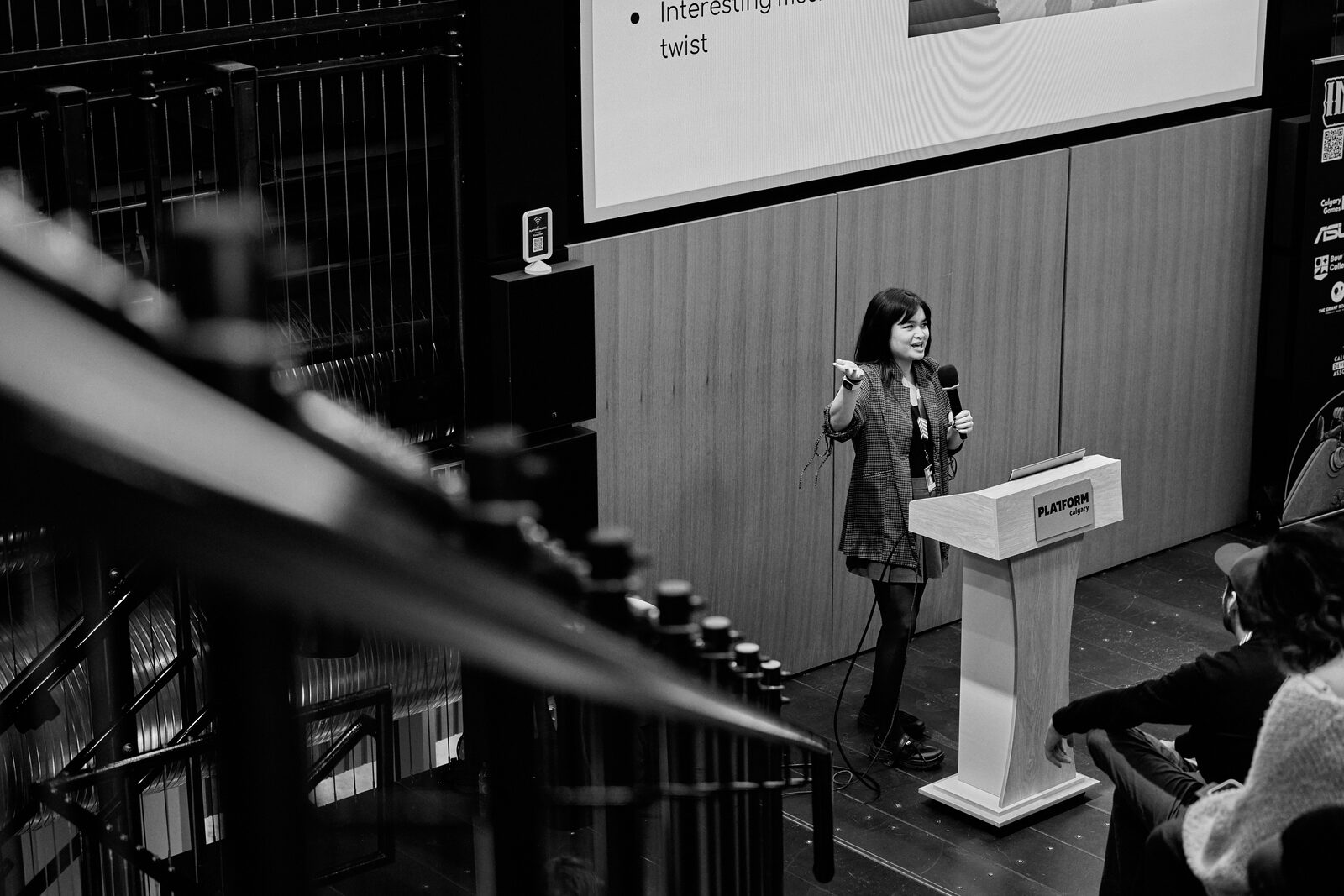 From the balcony, past the stage lighting rig: a speaker alone at the Platform podium with her slide behind her.