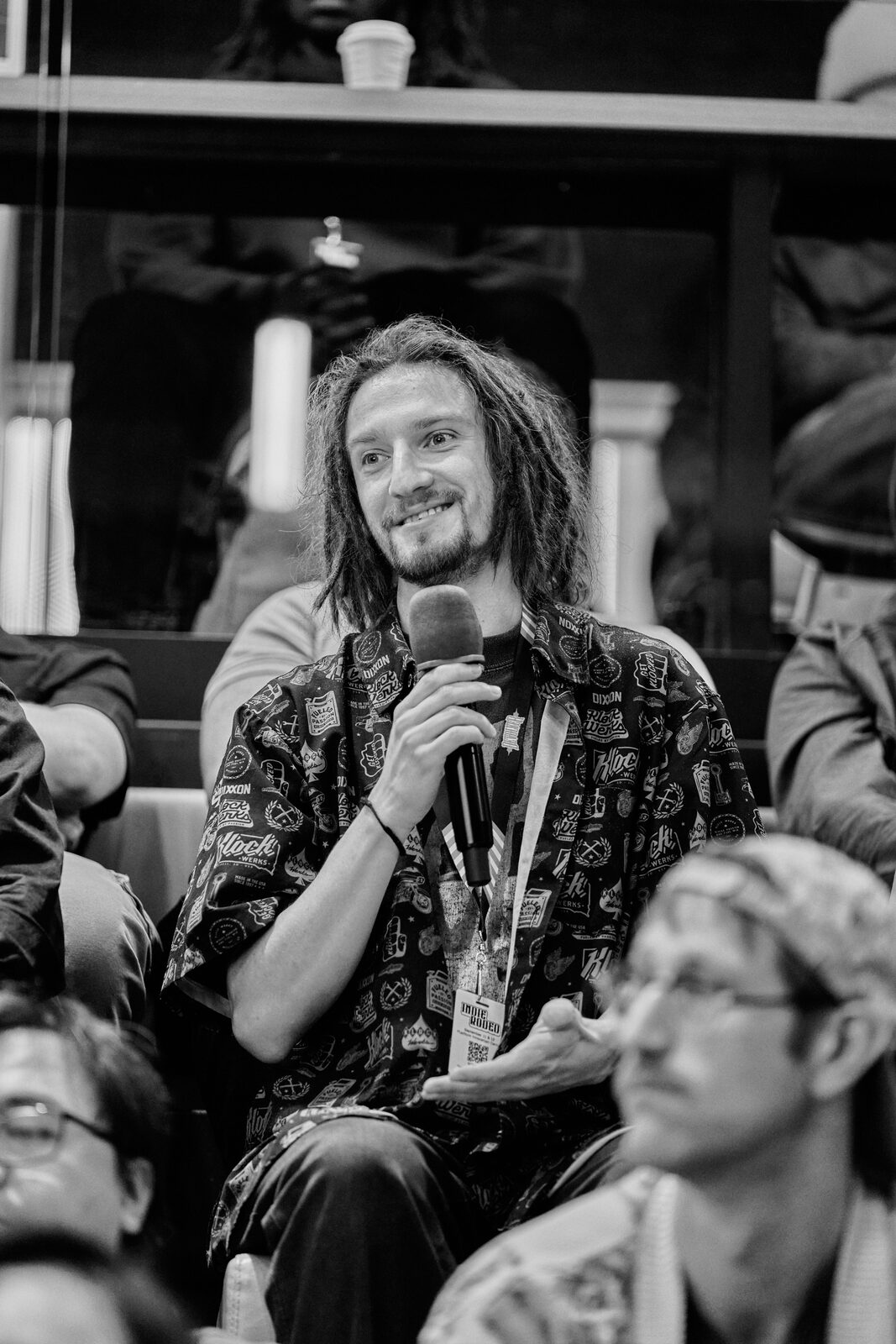 An attendee with long hair stands with the mic to ask a question, the seated rows around him listening.