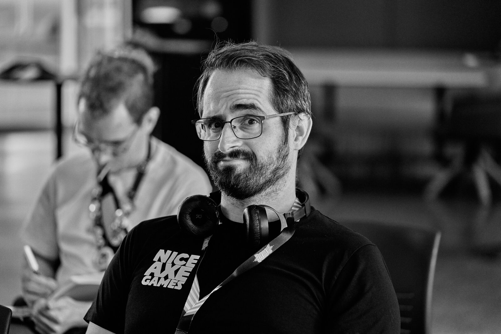 An attendee in a Nice Axe Games shirt with headphones round his neck looks toward the camera, a second attendee writing behind him.