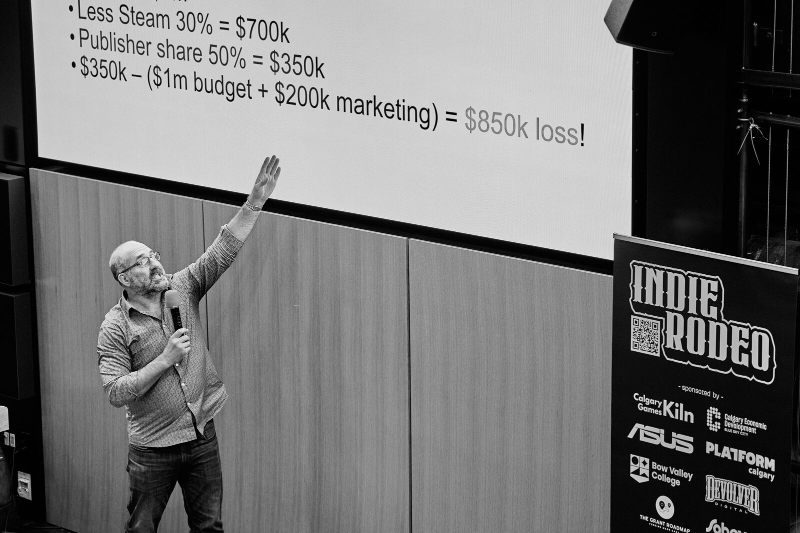 A speaker throws an arm toward a slide breaking down a publisher split, the arithmetic ending in an $850k loss, the Indie Rodeo sponsor banner beside the screen.