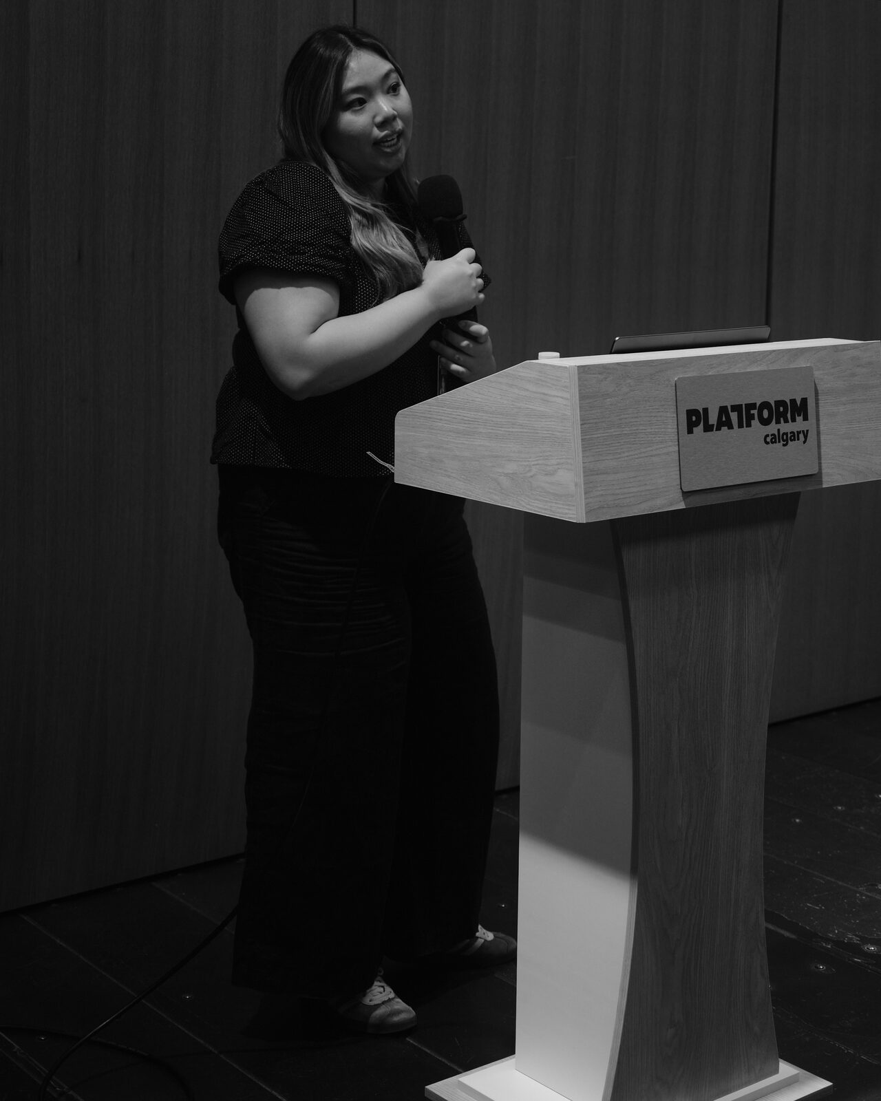 A speaker in a polka-dot top holds a mic beside the Platform Calgary podium, mid-sentence, lit against the dark wood-panel wall.