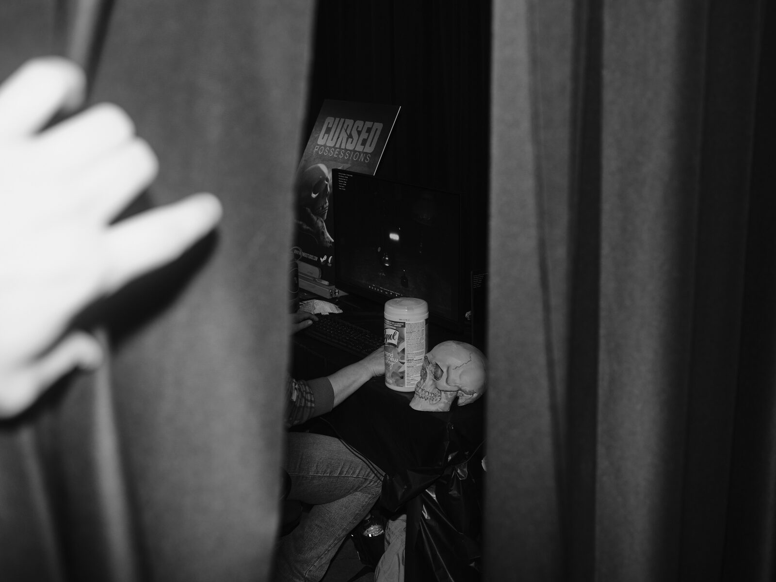 Peeking through a gap in black curtains: a player at the Cursed Possessions booth, a plastic skull and a canister of disinfecting wipes beside the keyboard, a hand holding the curtain open.