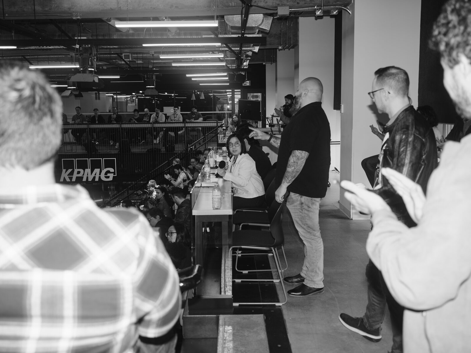 From the upper-level panel table, a seated attendee speaks into a mic while a tattooed attendee stands and points, others looking on, the KPMG sign across the atrium below.