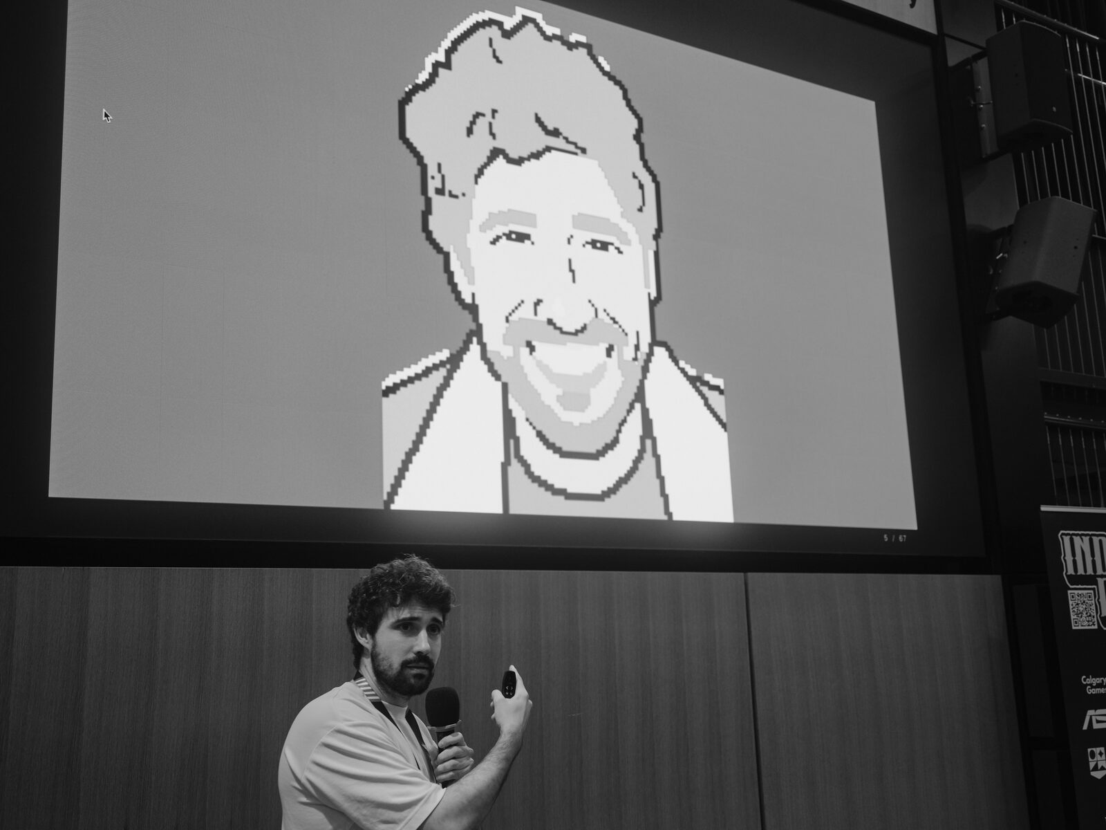 A curly-haired speaker holds a mic and clicker beneath a giant pixel-art self-portrait on the stage screen, an Indie Rodeo banner at the edge of frame.